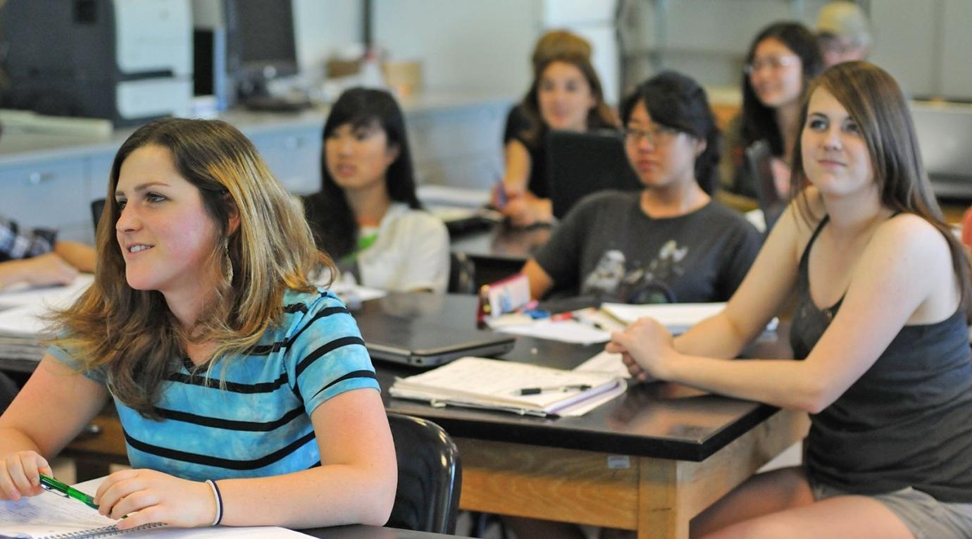 students sit at tables in a science classroom and watch the teacher lecture