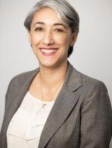 Woman with short gray hair, smiling, wearing gray blazer and white shirt, in front of gray background.