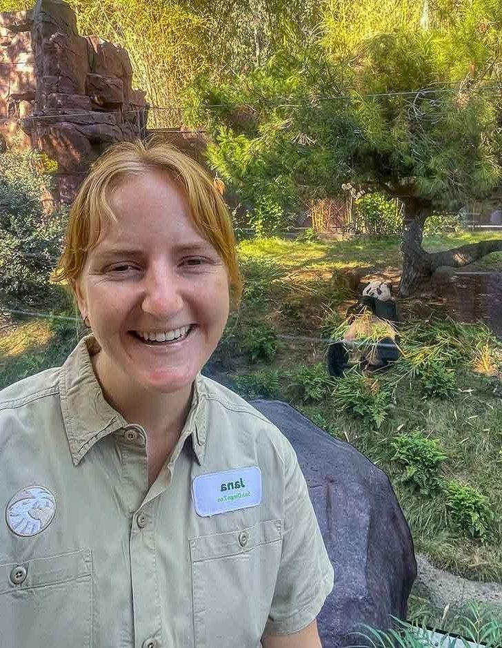 Jana Biedenwig smiles in the panda enclosure with a panda sitting behind her.