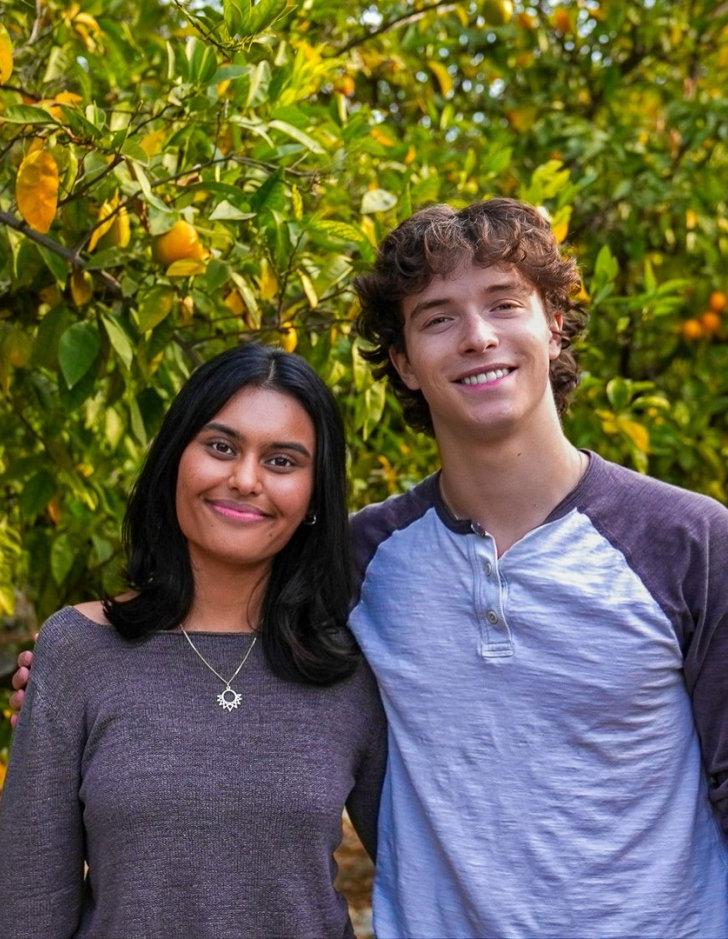 Sam McGinty and Aru Warrier smile and stand together in front of orange trees.