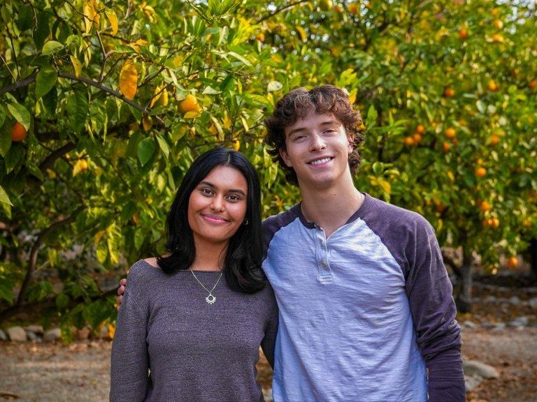 Sam McGinty and Aru Warrier smile and stand together in front of orange trees.